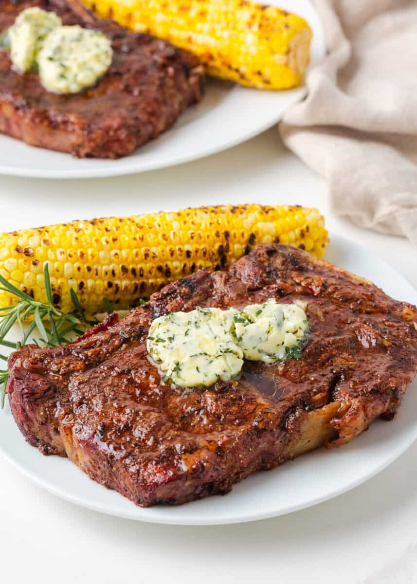 Perfect grilled steak topped with herb butter and served with grilled corn on a white plate.