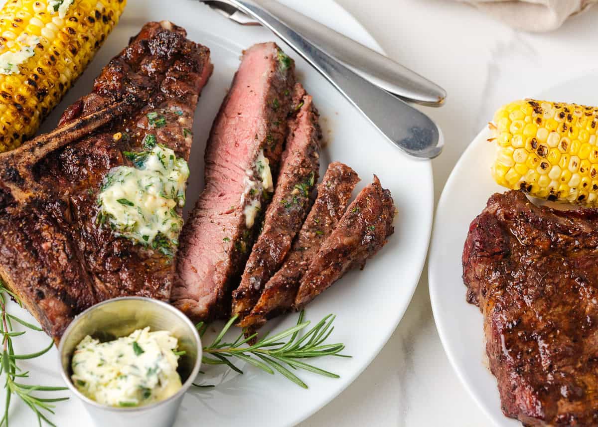 Grilled steak sliced on a white plate with herb butter, rosemary and corn for serving.