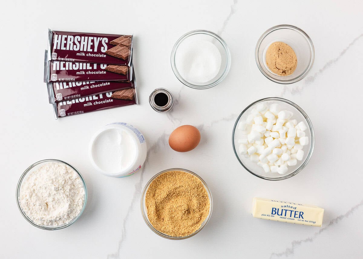 Ingredients for s’mores pie including Hershey bars, marshmallows, graham cracker crumbs, butter, egg, sugar, flour, vanilla, and marshmallow creme.