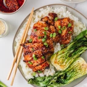Grilled Asian chicken thighs on a shallow plate with white rice with bok choy, sesame seeds, green onions and chili crunch sauce.