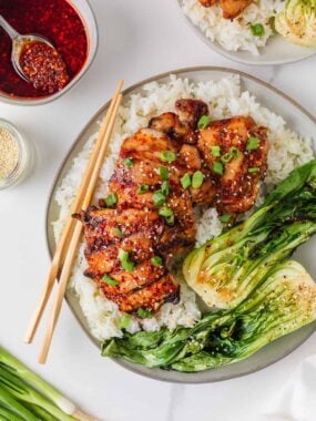 Grilled Asian chicken thighs on a shallow plate with white rice with bok choy, sesame seeds, green onions and chili crunch sauce.