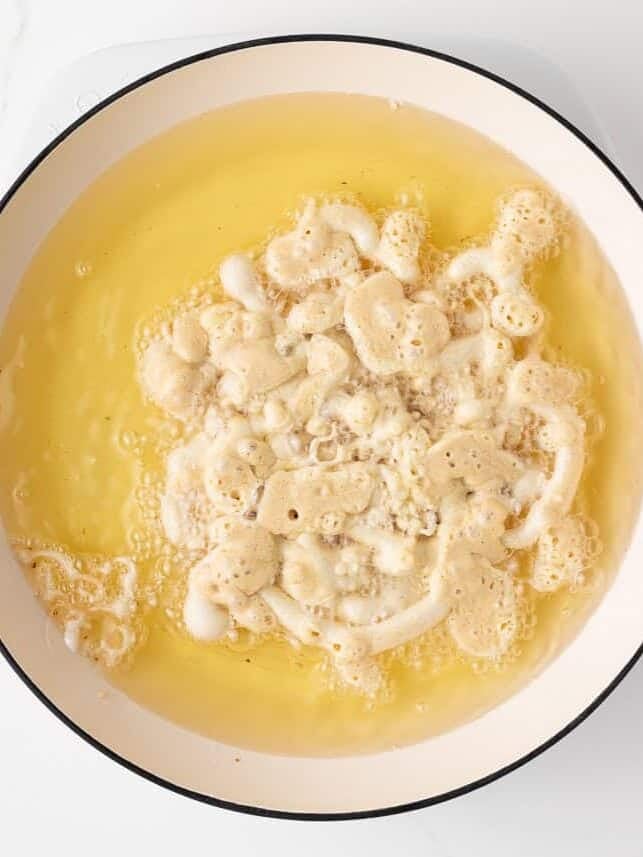 Pale funnel cake batter just hitting the hot oil in a white Dutch oven, bubbling around the spirals.