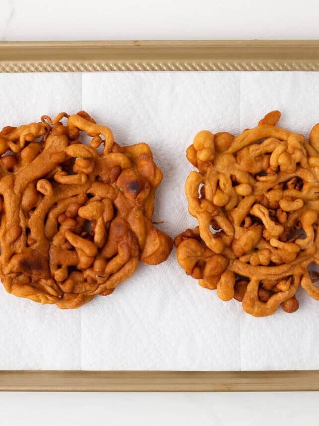 Two golden brown homemade funnel cakes draining on a paper towel-lined sheet pan.