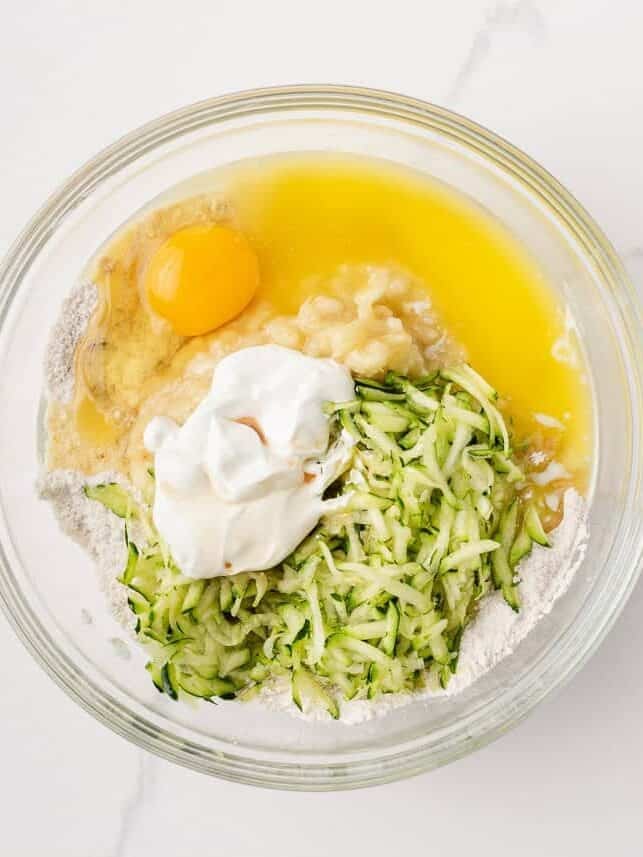 Moist, tender, and packed with flavor, this zucchini banana bread is a delicious way to use up ripe bananas and fresh zucchini in one easy loaf! Zucchini banana bread ingredients including grated zucchini, mashed banana, egg, sour cream and melted butter in a glass bowl.
