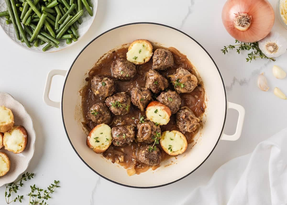 Skillet of French onion meatballs in beef gravy topped with melted cheese baguette slices.