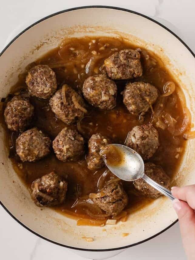 Cooked meatballs simmering in French onion gravy with a spoon basting the sauce over the top.