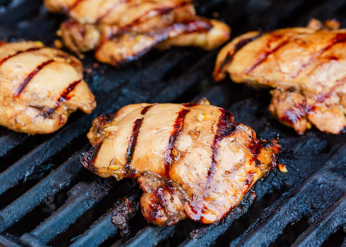 Marinated Asian chicken thighs cooking on a hot grill with charred grill marks.