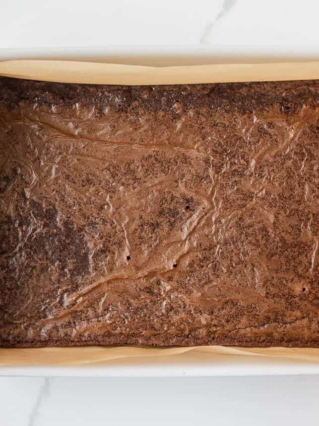 Baked brownie layer cooling in a parchment-lined pan before adding the peanut butter topping.