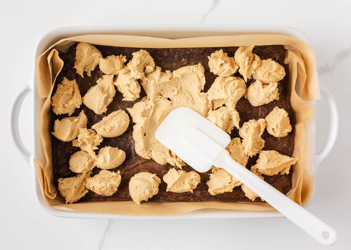 Peanut butter layer being spread over cooled brownies in a parchment-lined baking dish.