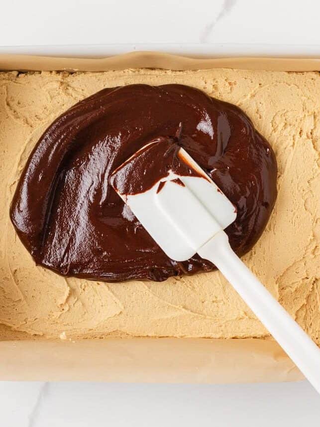 Chocolate ganache being spread over the peanut butter layer in a buckeye brownie recipe.