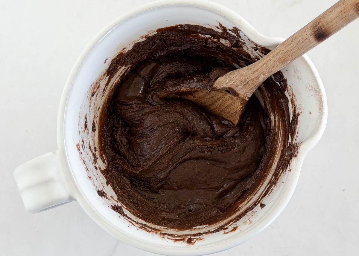 Thick chocolate cake mix cookie dough being stirred in a white mixing bowl with a wooden spoon.