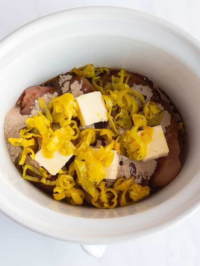 Raw chicken breasts in a white crockpot topped with ranch seasoning, au jus mix, pats of butter, and sliced pepperoncini peppers before cooking.