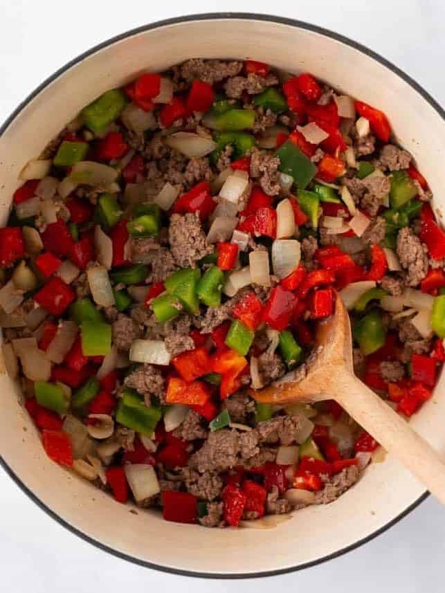 Browned ground beef with chopped onion and red and green bell peppers cooking in a white Dutch oven, a wooden spoon resting in the pot.