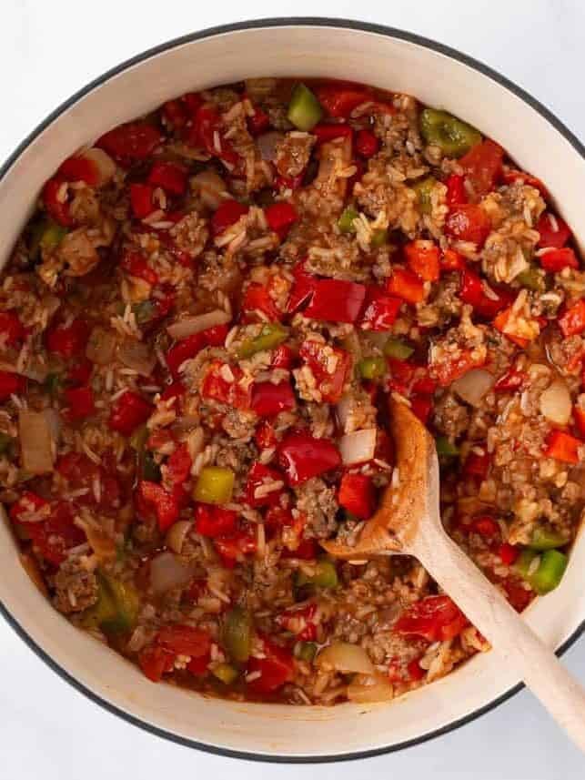 Stuffed pepper casserole after simmering, with tender rice, ground beef and bell peppers in a thick tomato sauce, a wooden spoon in the white pot.