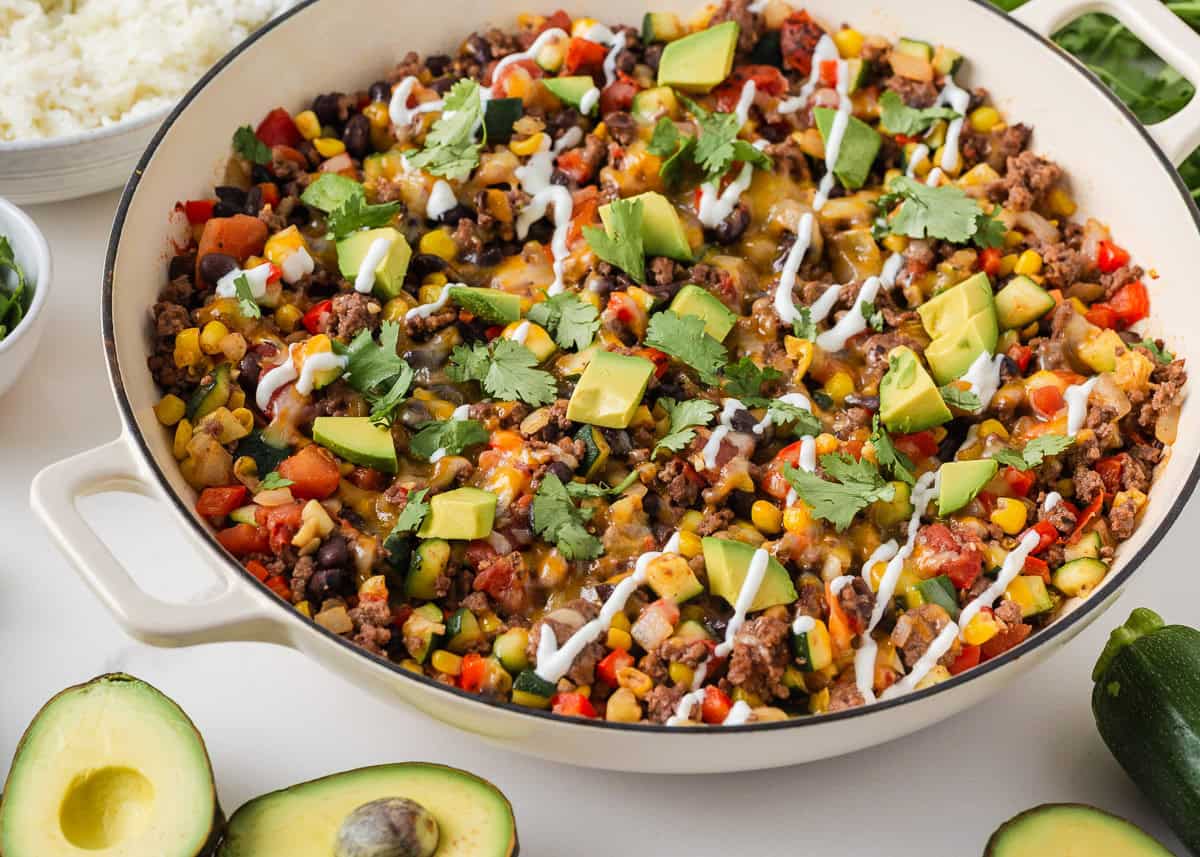 Ground beef and zucchini skillet recipe served in a large pan with avocado, cilantro and sour cream on top.
