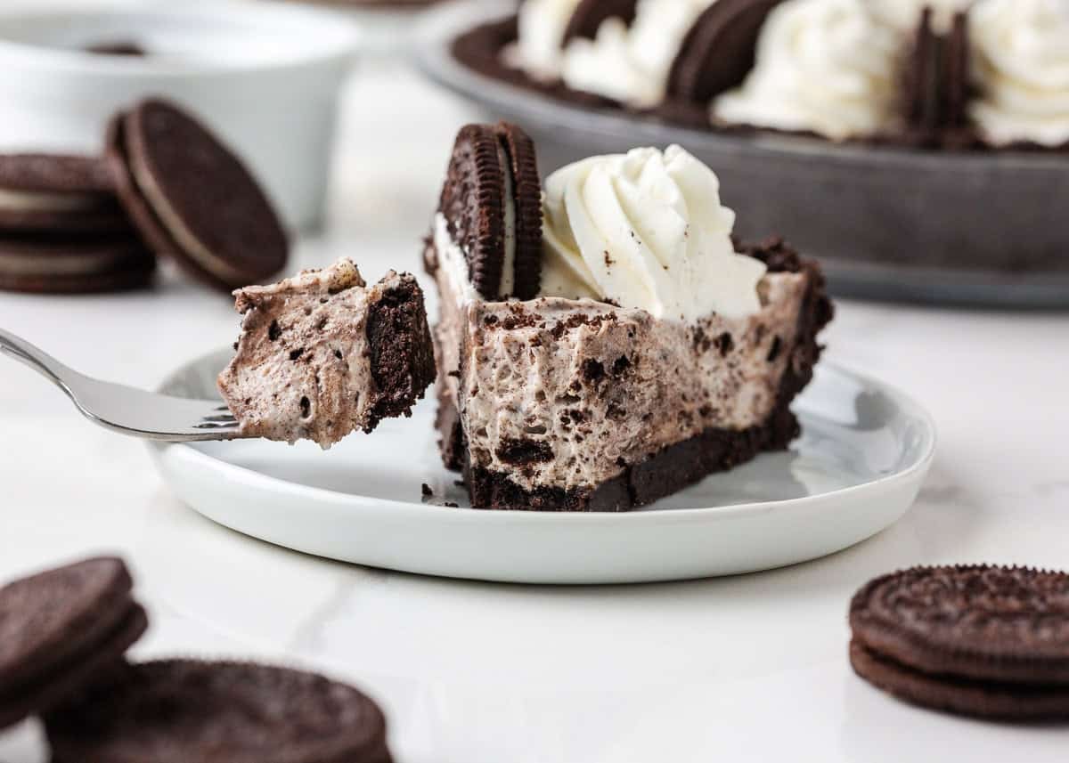Slice of no bake Oreo cheesecake on a plate with whipped cream and a halved Oreo, with a bite on a fork.