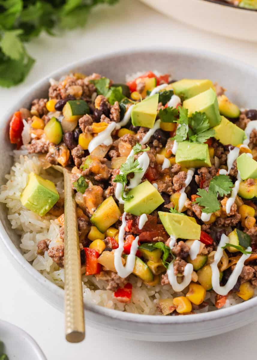 Bowl of ground beef and zucchini skillet served over rice with avocado, cilantro and a drizzle of sour cream.