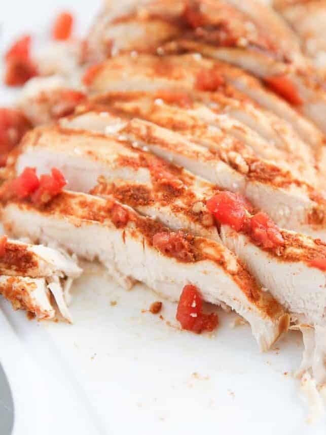 Sliced cooked chicken breast seasoned with taco seasoning and diced tomatoes on a cutting board.