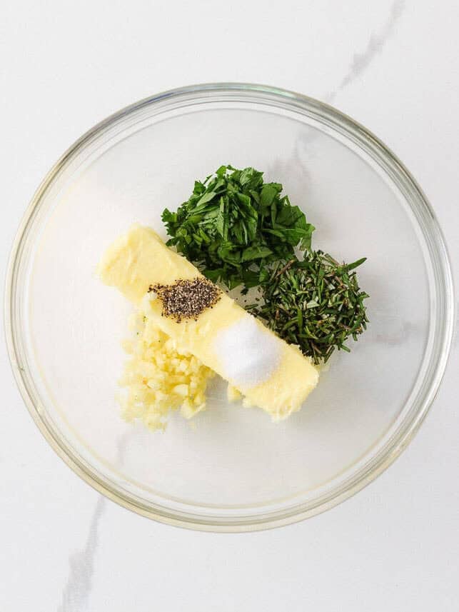 Softened butter in a bowl with garlic, rosemary, parsley, salt and pepper for herb butter.