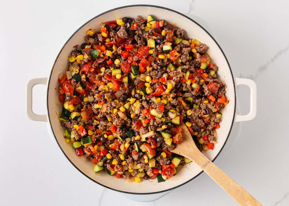 Cooked ground beef and zucchini dinner being stirred together with black beans, corn, tomatoes, onion and red bell pepper in a skillet.