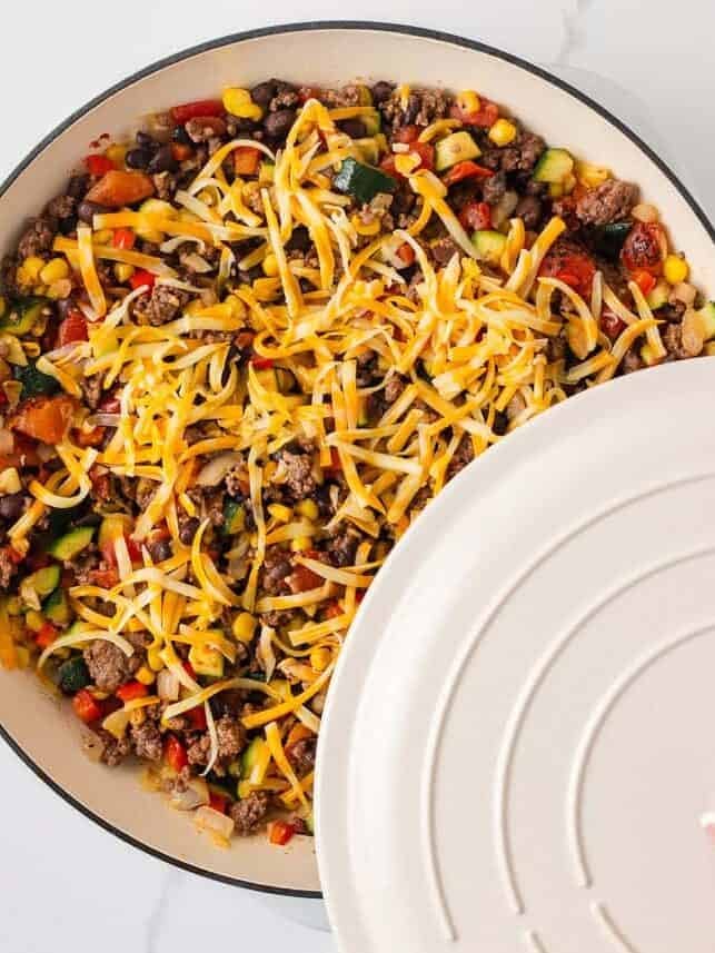 Shredded cheese being added to a ground beef and zucchini skillet before covering the pan to melt.
