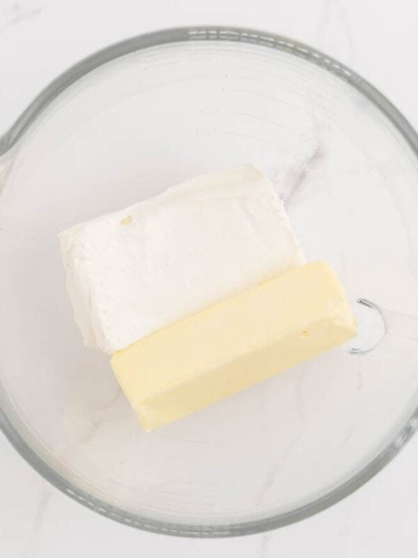A block of cream cheese and a stick of softened butter in a glass mixing bowl.