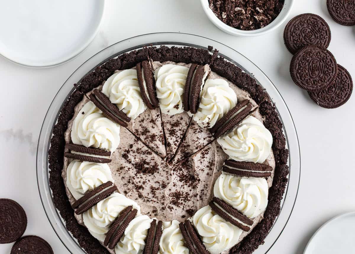 Sliced no bake Oreo cheesecake in an Oreo crust topped with whipped cream swirls, halved Oreos and cookie crumbs.