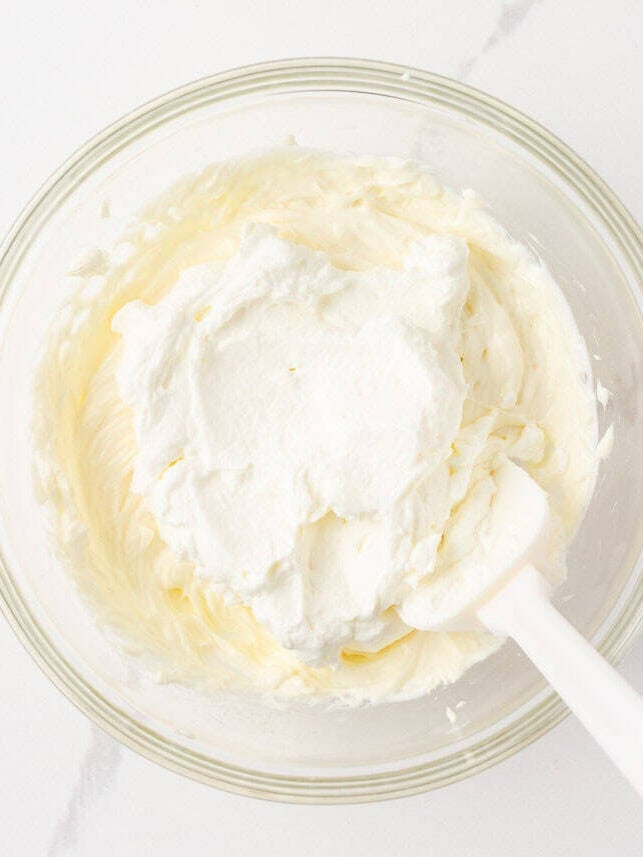 Whipped cream added to the cream cheese mixture in a glass bowl before folding together.