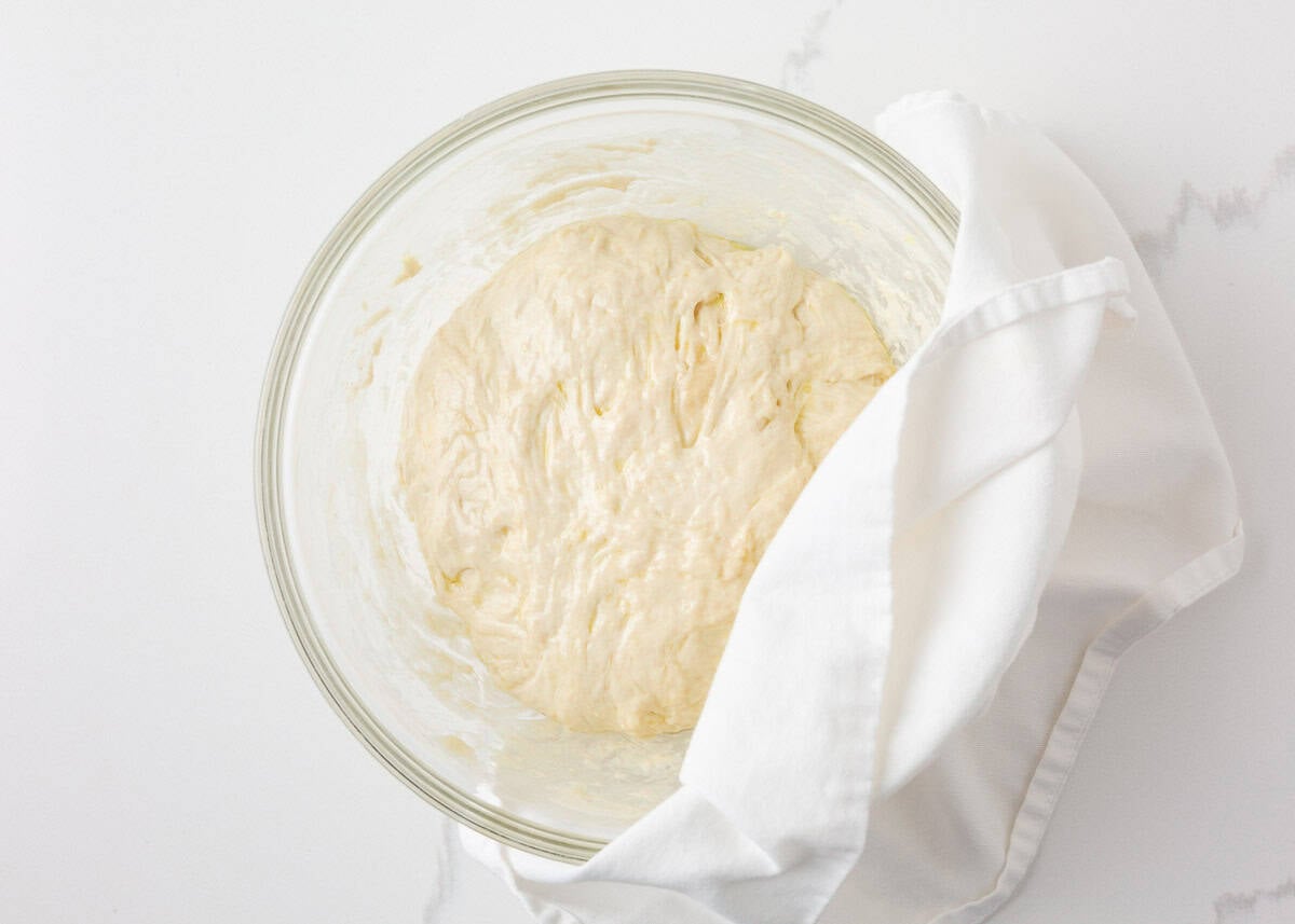 Sourdough French bread dough rising in a glass bowl covered with a white kitchen towel.