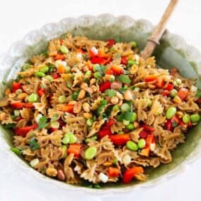 Thai pasta salad with bow tie pasta, red bell pepper, edamame, peanuts, and cilantro in a green bowl with a wooden spoon.