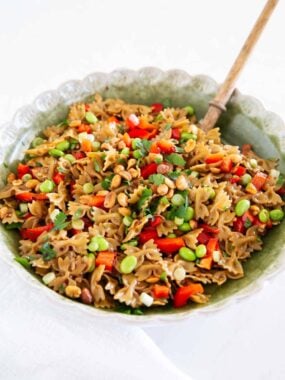 Thai pasta salad with bow tie pasta, red bell pepper, edamame, peanuts, and cilantro in a green bowl with a wooden spoon.
