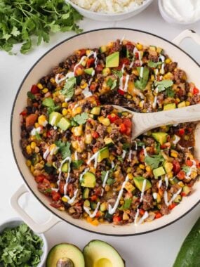 Zucchini beef skillet topped with melted cheese, avocado, cilantro and sour cream in a large cream-colored pan.