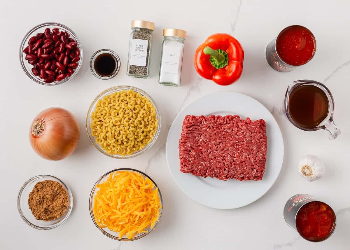 Chili mac ingredients arranged on a countertop, including ground beef, elbow pasta, kidney beans, tomatoes, cheddar cheese, bell pepper, onion and seasonings.