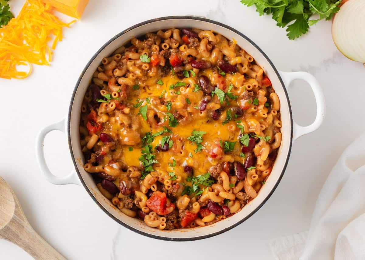 Overhead view of homemade chili mac with ground beef, kidney beans, elbow pasta, tomatoes and melted cheddar cheese in a white Dutch oven.