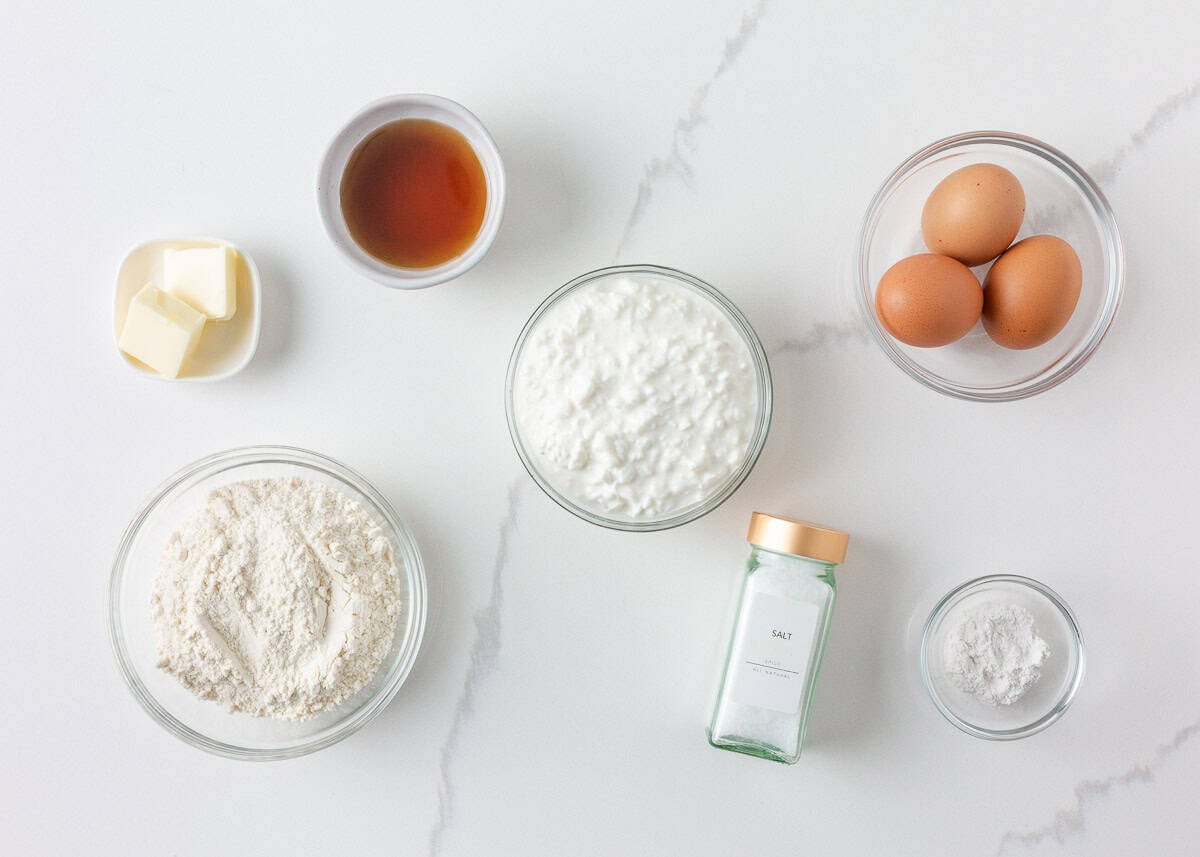 Cottage cheese pancake ingredients arranged in bowls on a white marble surface, including cottage cheese, eggs, flour, maple syrup, butter, baking powder and salt.