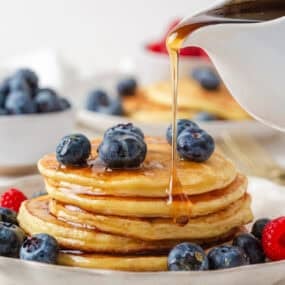Maple syrup pouring from a white pitcher over a stack of golden cottage cheese pancakes topped with blueberries and served with fresh berries.