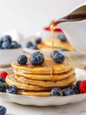 Maple syrup pouring from a white pitcher over a stack of golden cottage cheese pancakes topped with blueberries and served with fresh berries.