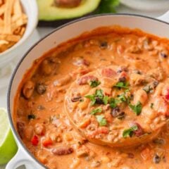 Close-up of creamy taco soup in a Dutch oven with a full ladle, fresh cilantro, tortilla strips and lime wedges nearby.