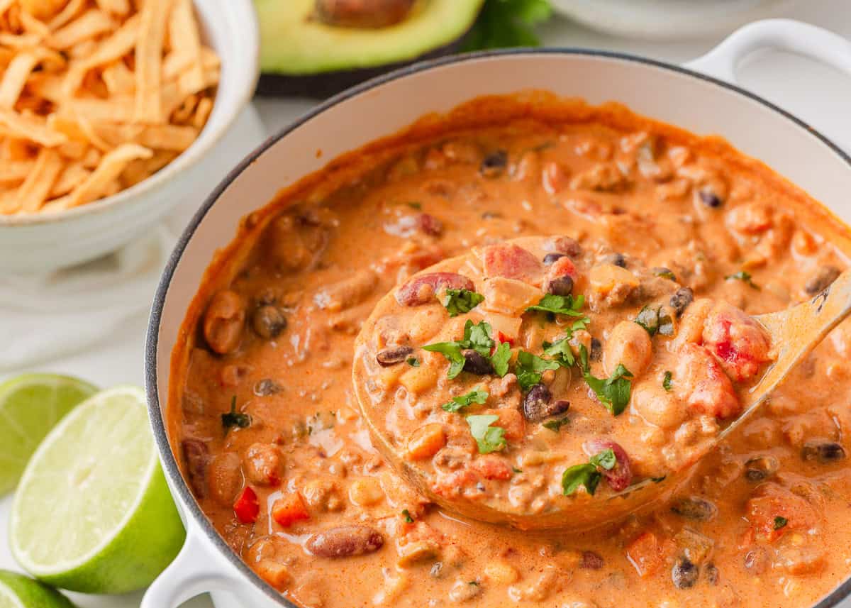Close-up of creamy taco soup in a Dutch oven with a full ladle, fresh cilantro, tortilla strips and lime wedges nearby.