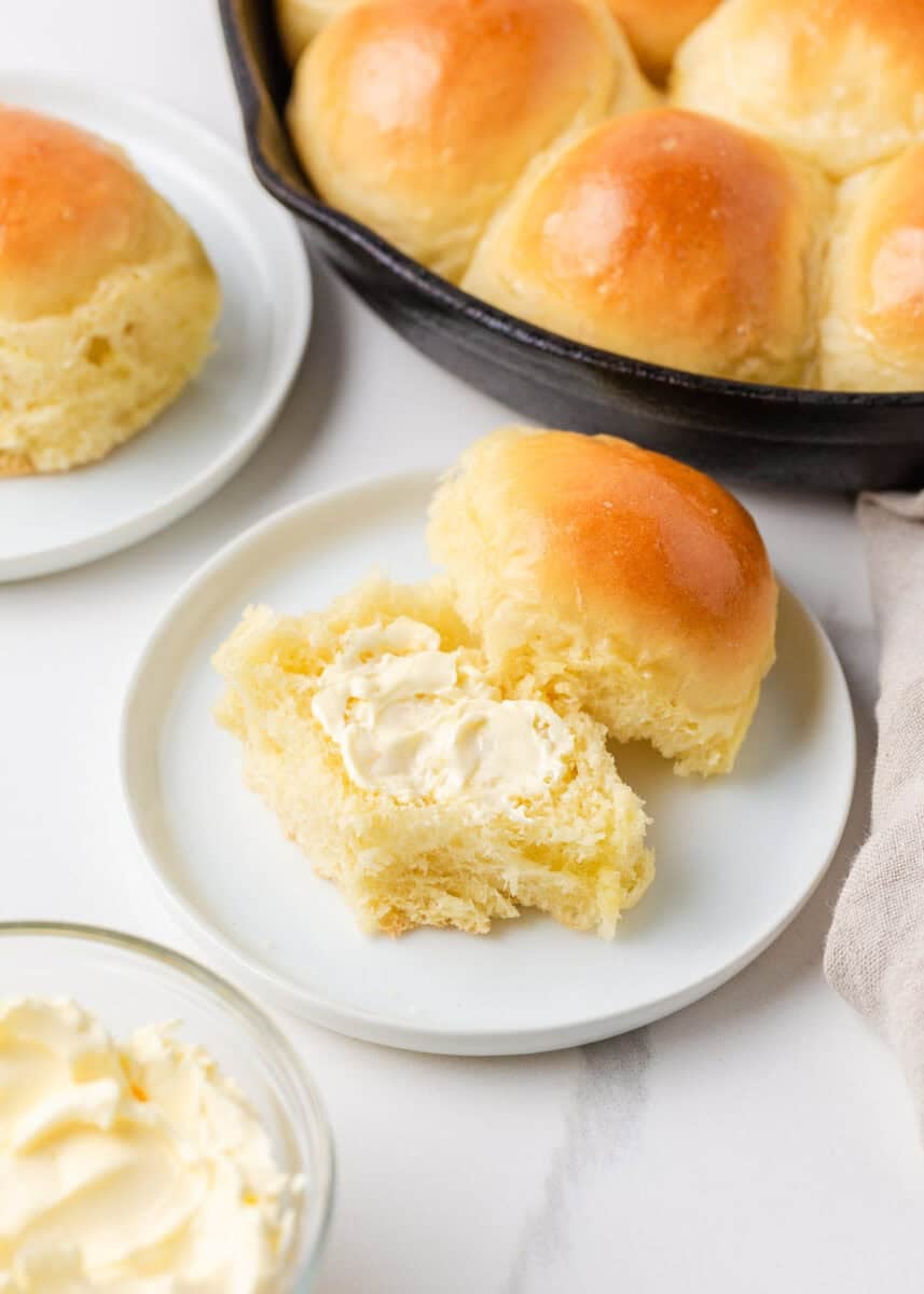 Soft skillet dinner roll torn open and spread with butter beside a cast iron pan of golden rolls.