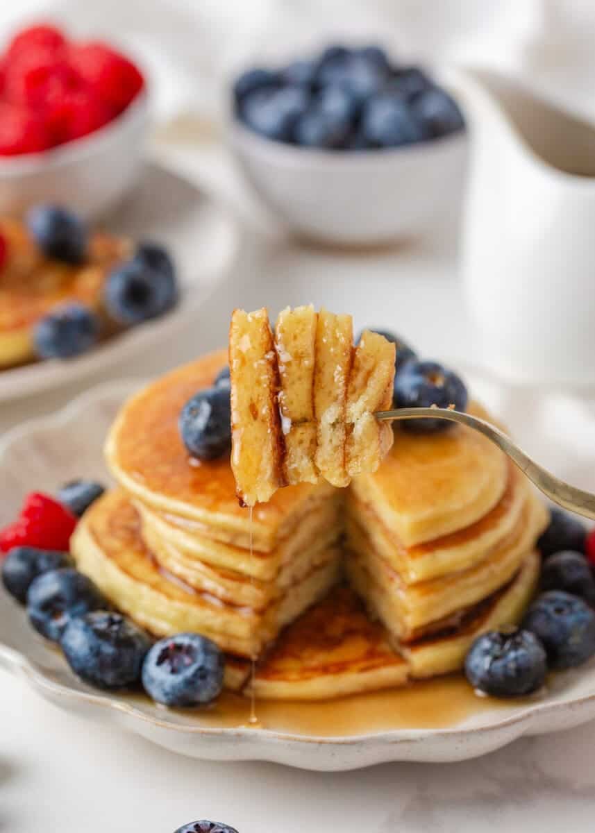 Fork holding a syrup-covered bite above a sliced stack of fluffy cottage cheese pancakes surrounded by fresh blueberries and raspberries.