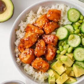 Teriyaki salmon rice bowl topped with cucumber, edamame, avocado, green onions and sesame seeds.