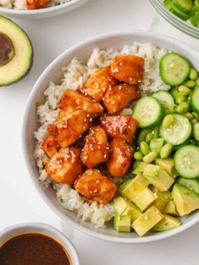Teriyaki salmon rice bowl topped with cucumber, edamame, avocado, green onions and sesame seeds.