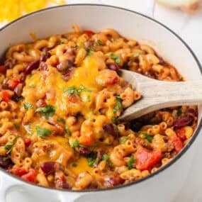 Chili mac with ground beef, kidney beans and melted cheddar cheese being served from a white Dutch oven with a wooden spoon.
