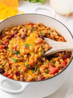 Chili mac with ground beef, kidney beans and melted cheddar cheese being served from a white Dutch oven with a wooden spoon.