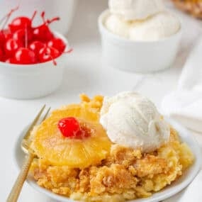 Pineapple dump cake served on a white plate with a pineapple ring, cherry, and scoop of vanilla ice cream.