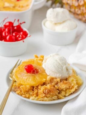 Pineapple dump cake served on a white plate with a pineapple ring, cherry, and scoop of vanilla ice cream.