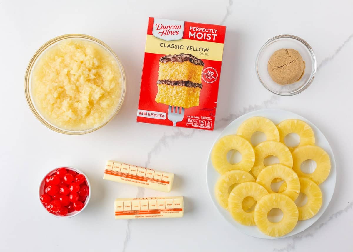 Ingredients for pineapple dump cake including crushed pineapple, yellow cake mix, butter, pineapple rings, cherries, and brown sugar.