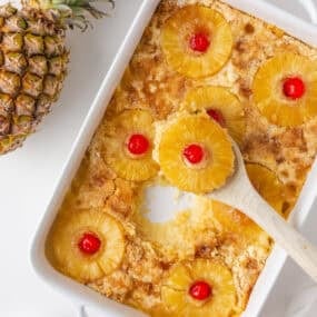 Overhead view of baked pineapple dump cake in a white casserole dish with pineapple rings, cherries, and a serving spoon.
