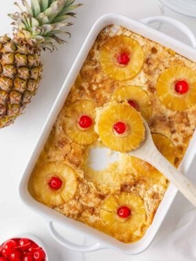 Overhead view of baked pineapple dump cake in a white casserole dish with pineapple rings, cherries, and a serving spoon.